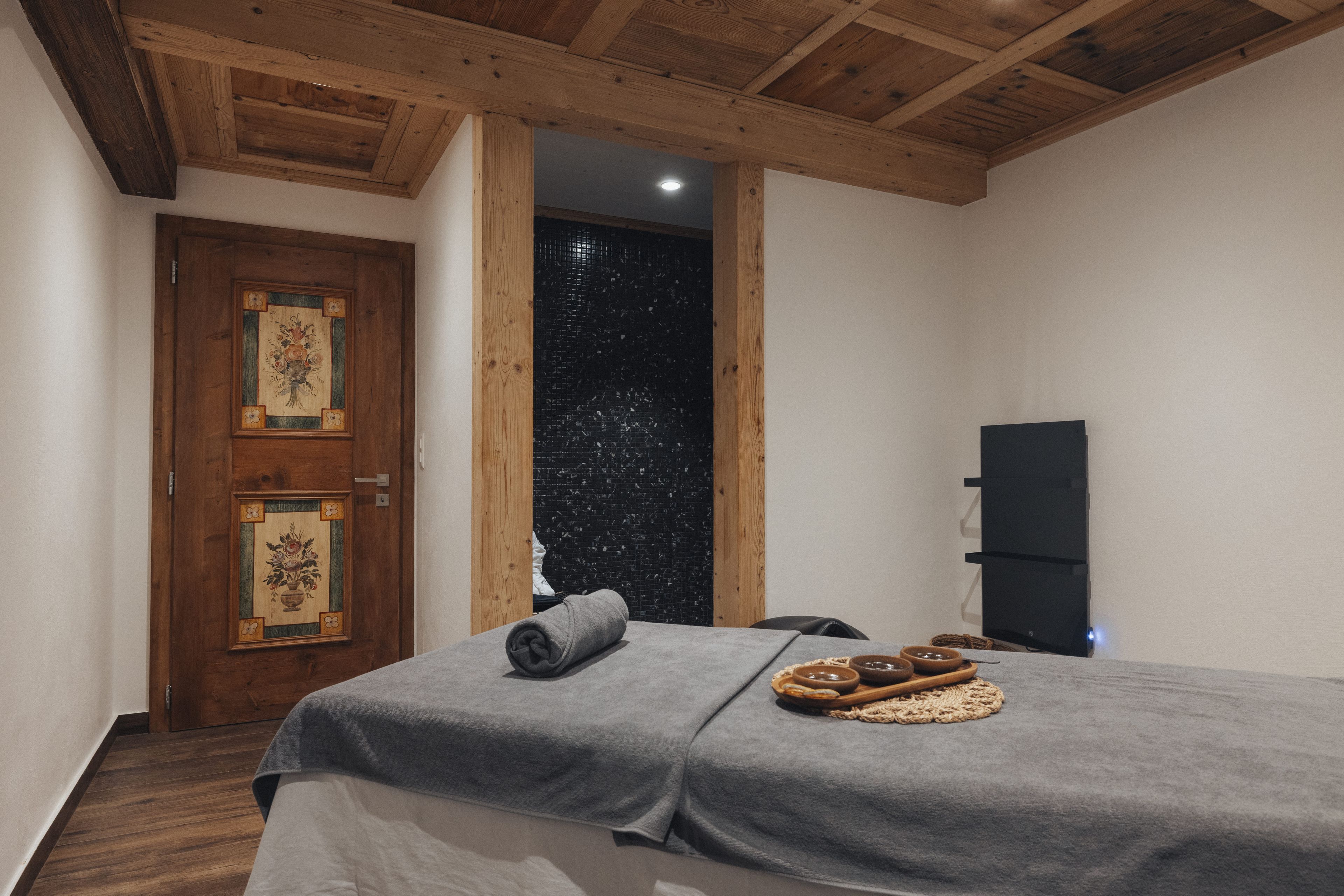 Ein Zimmer mit einem Bett, einer Holztür mit floralem Dekor, einer schwarzen Fliesenwand und einer Decke mit Holzbalken.