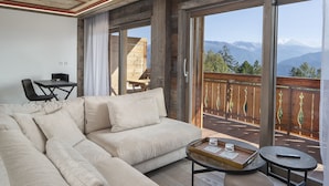 A modern living room with a large beige sectional sofa, a small round table with a tray, and a view of mountains through large windows. - Aïda Hôtel & Spa (Adults Only) (Crans-Montana)