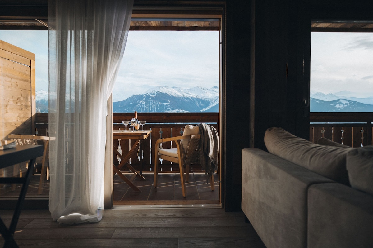 Une pièce avec un plancher en bois, une table entourée de chaises, un canapé et une vue sur des montagnes enneigées, visible à travers une porte vitrée.