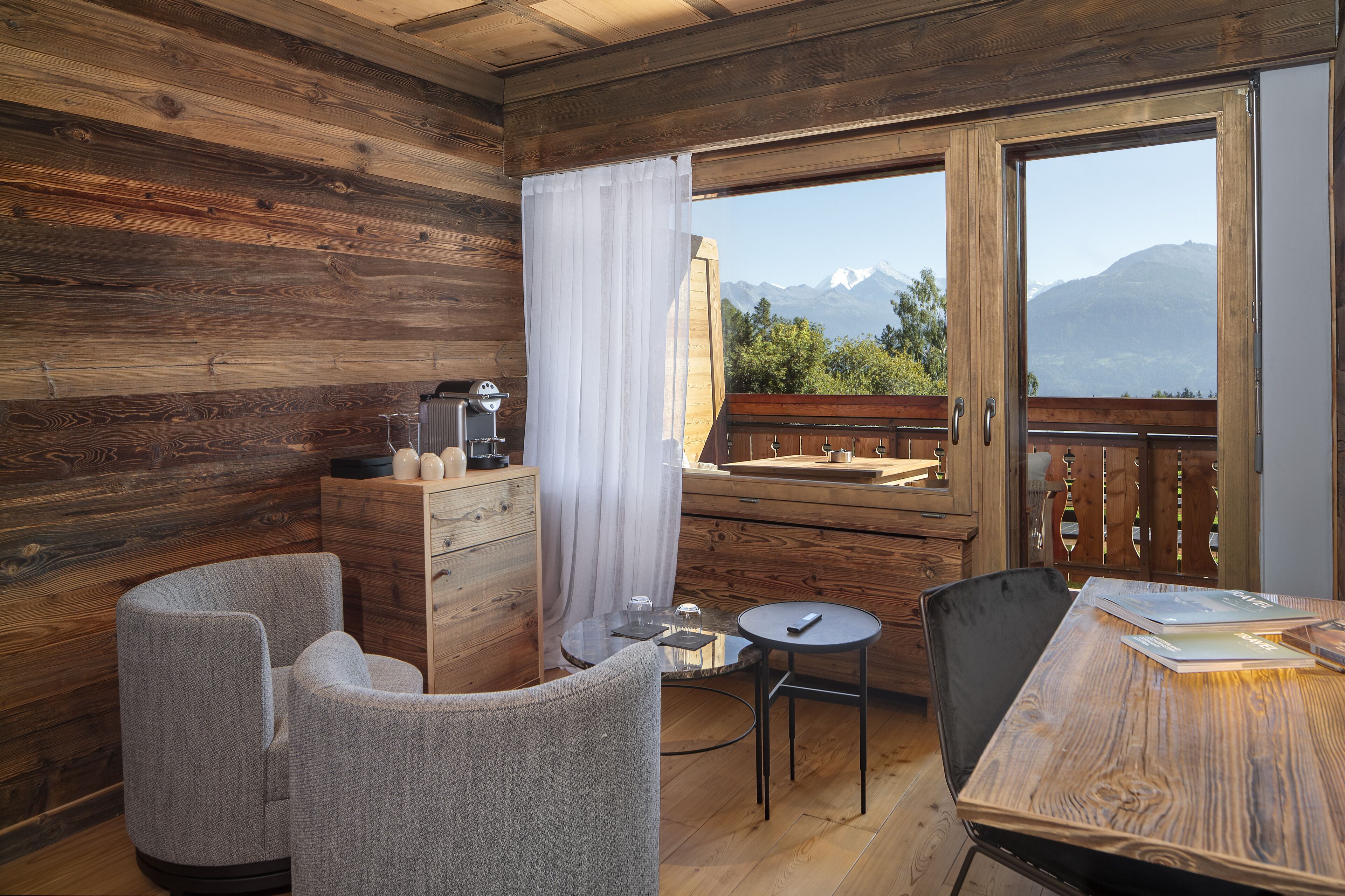 Una cabaña de madera con un balcón, una mesa de comedor, dos sillas, una mesita con una lámpara y vistas a las montañas.