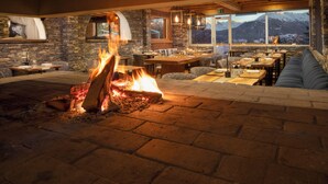 A cozy interior with a stone fireplace, wooden furniture, and a view of mountains through large windows. - Aïda Hôtel & Spa (Adults Only) (Crans-Montana)