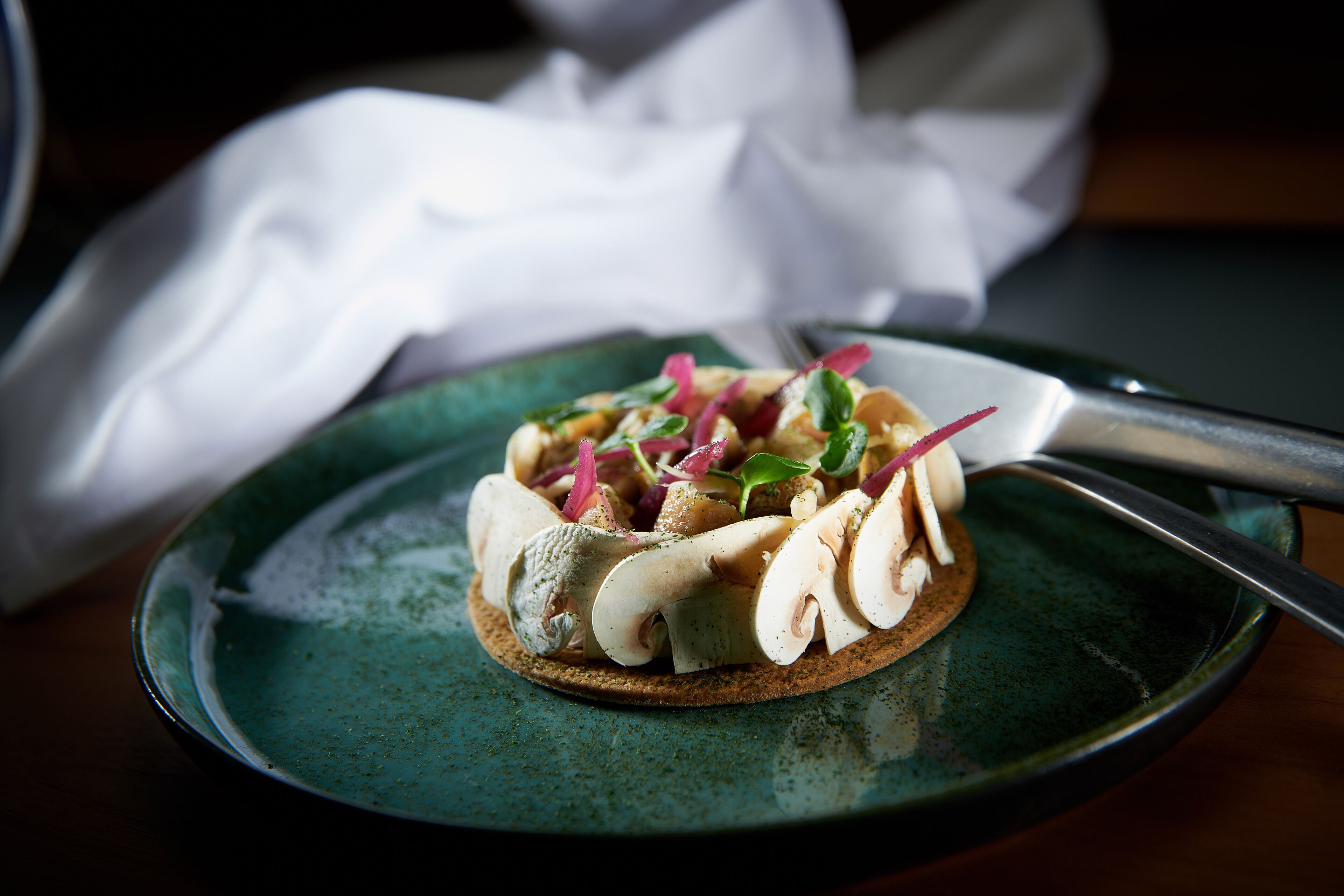 Une assiette avec un cracker garni d’une tranche de champignon, un couteau et une serviette.
