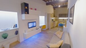 Ein modernes Wohnzimmer mit einem Flachbildfernseher, einer Couch und einem Essbereich, im Hintergrund ist eine Küche zu sehen.