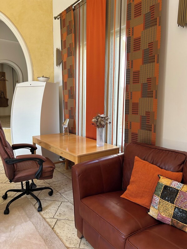 A room with a brown leather sofa, a wooden desk, a brown office chair, and a patterned curtain.