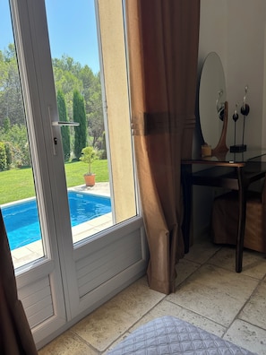 A room with a sliding glass door leading to a pool, a wooden desk with a mirror, and a small potted plant.