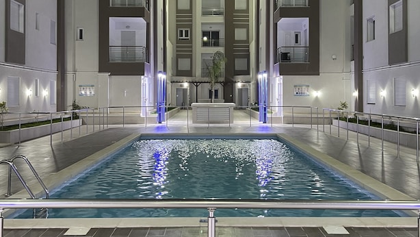 Vista nocturna de un hotel moderno con piscina central, rodeado de edificios de varios pisos con balcones y ventanas iluminadas.