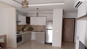 Una cocina moderna con armarios blancos, horno empotrado y refrigerador. Se observa una puerta de madera y una pequeña mesa de madera.