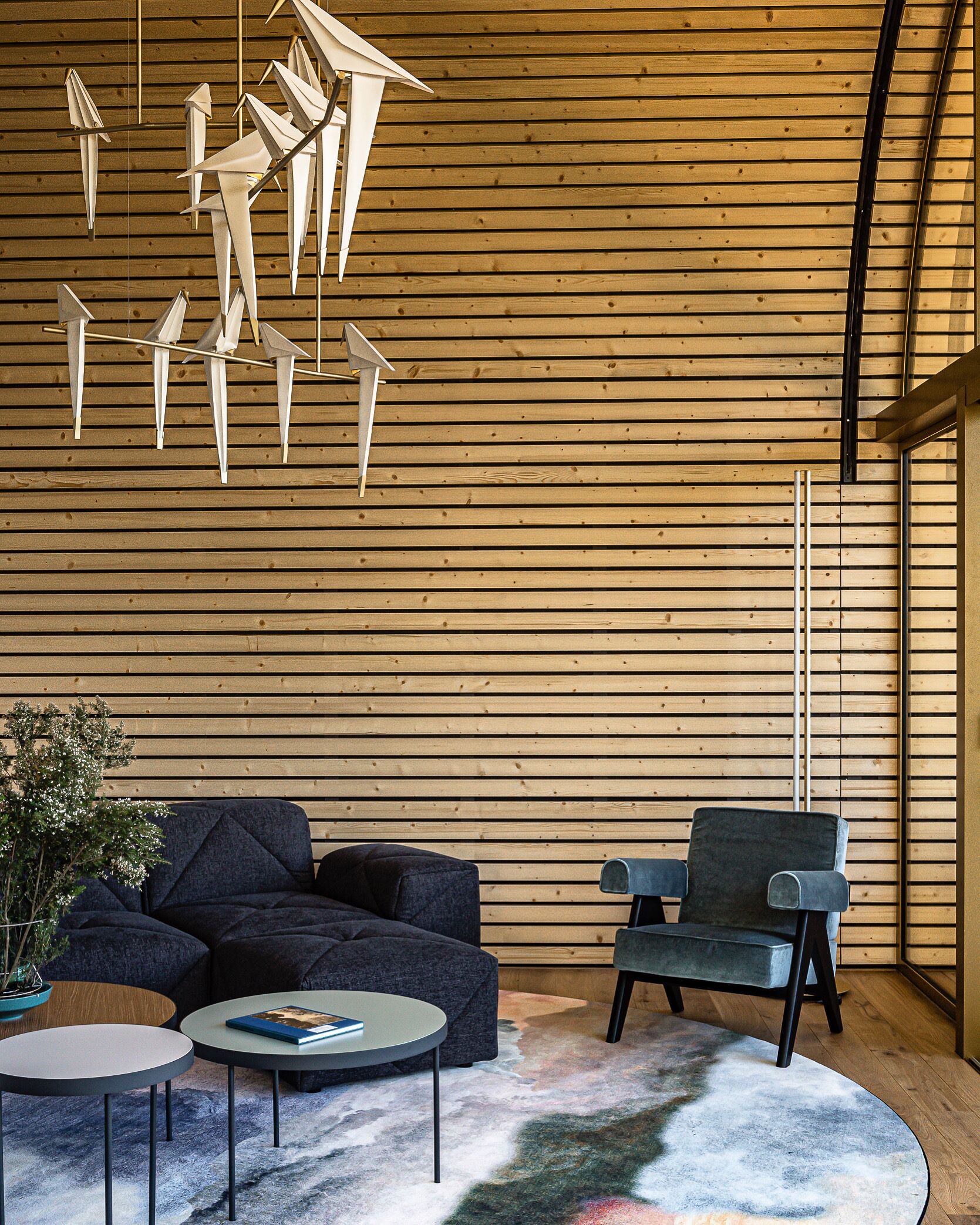 A modern interior with a wooden slatted ceiling, a dark sofa, a single armchair, and a small round table with a book.