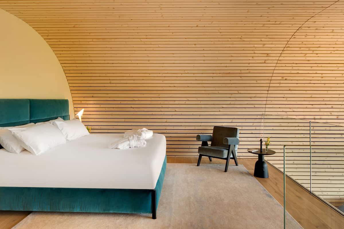 A modern bedroom with a curved ceiling, a teal bed, white bedding, a small round table with a vase, and a chair.