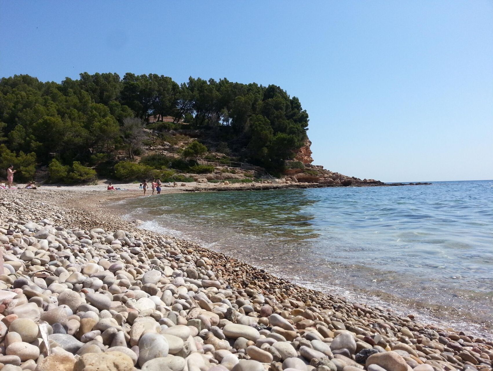 A pebbled beach with clear water, a rocky cliff with trees, and a few people in the distance.
