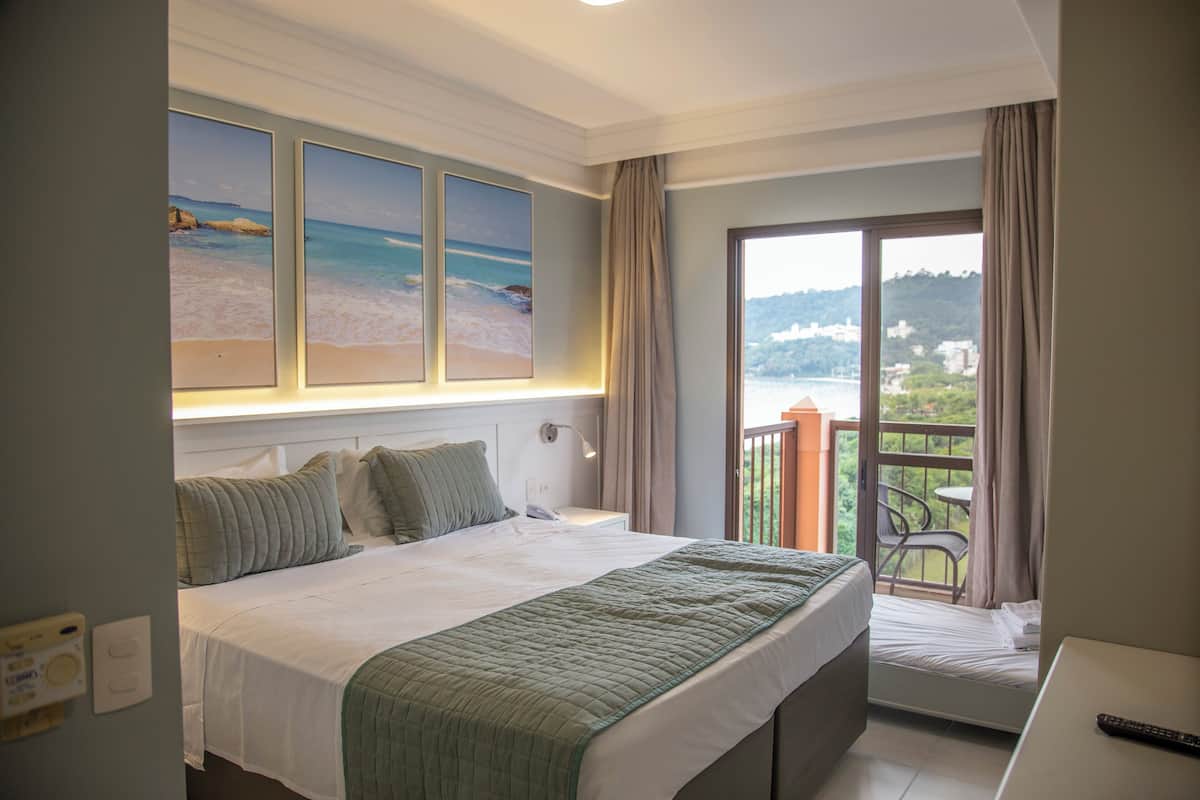 A hotel room with a bed, a desk, and a balcony with a view of the sea and buildings.