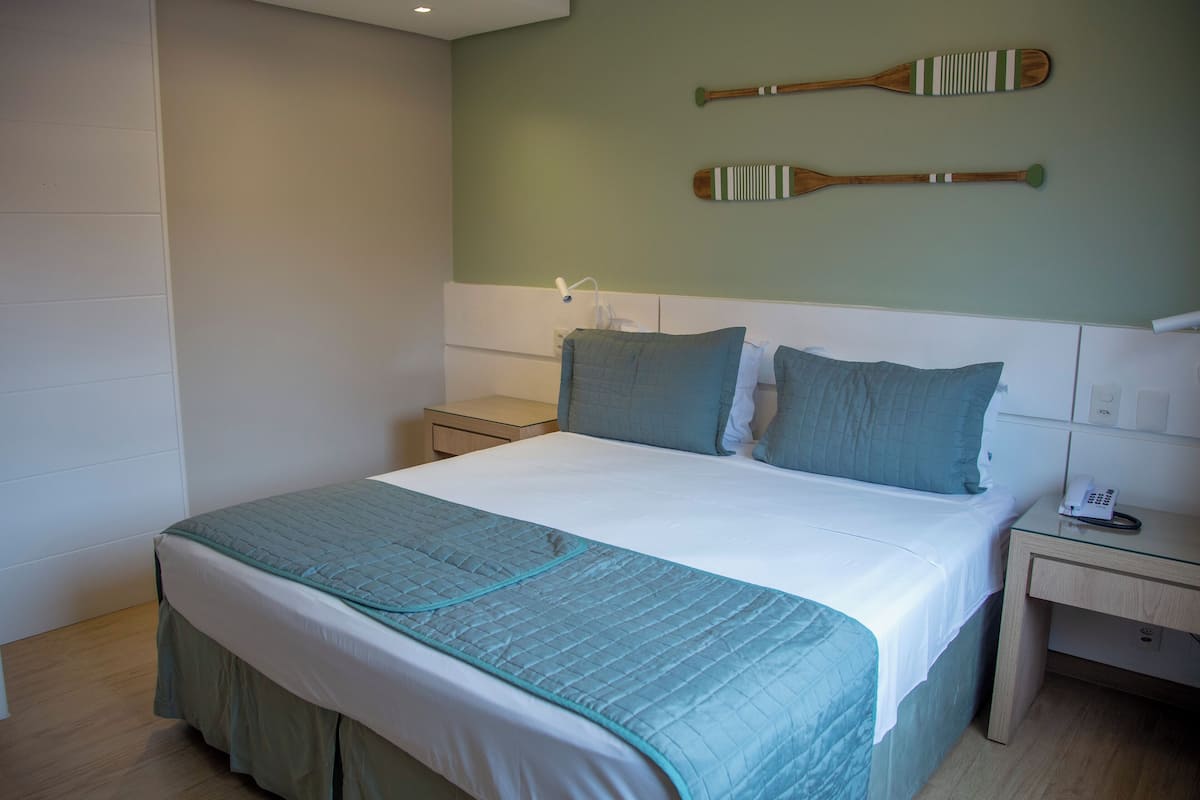 A neatly made bed with a teal bedspread and pillows, a bedside table with a phone, and two wooden paddles on the wall.
