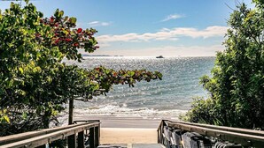 Une passerelle en bois mène à une plage d’où l’on a une vue dégagée sur l’océan et où l’on aperçoit un bateau au loin.