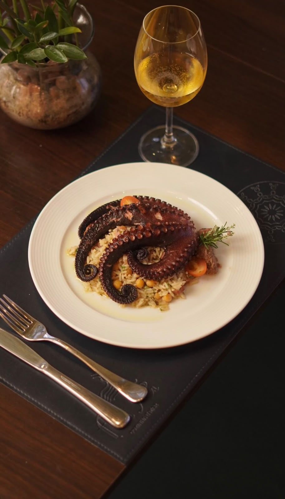Un plato de pulpo con arroz y verduras, una copa de vino blanco y un tenedor sobre un camino de mesa oscuro.