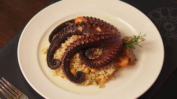 Un plato de pulpo con arroz y verduras, una copa de vino blanco y un tenedor sobre un camino de mesa oscuro.