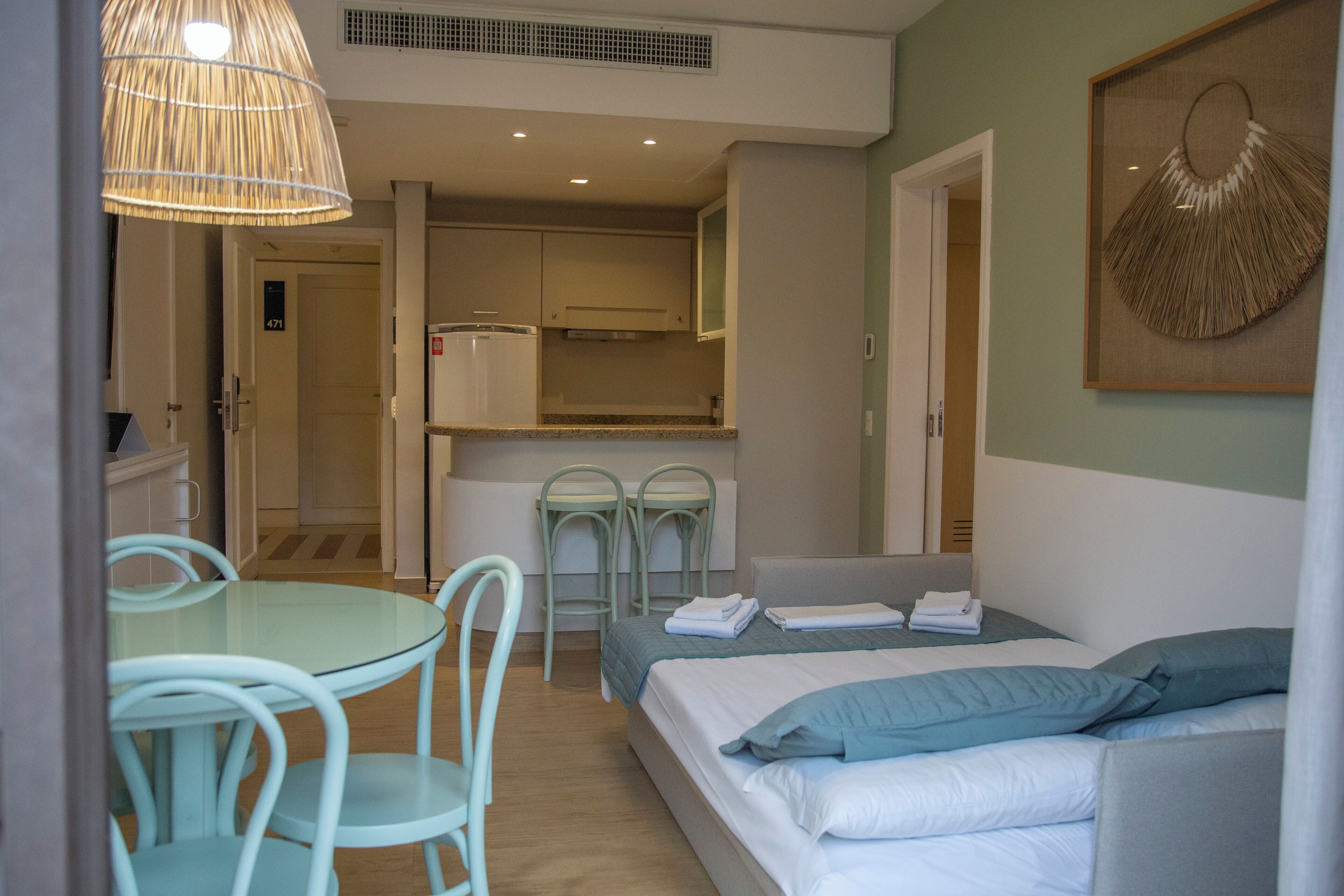Une chambre d’hôtel compacte comprenant un lit, une petite kitchenette, un coin repas avec une table et des chaises, et un luminaire suspendu.