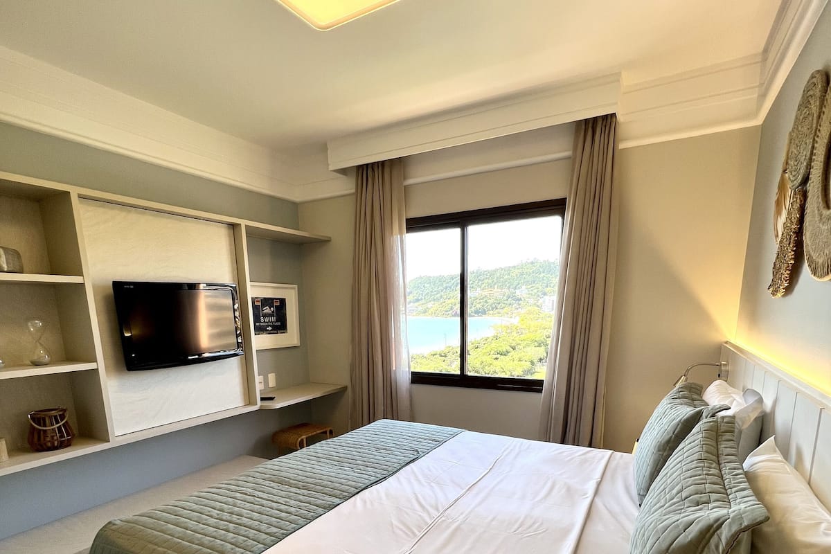 A hotel room with a bed, a flat-screen TV on a shelf, a window with a view, and a ceiling light.