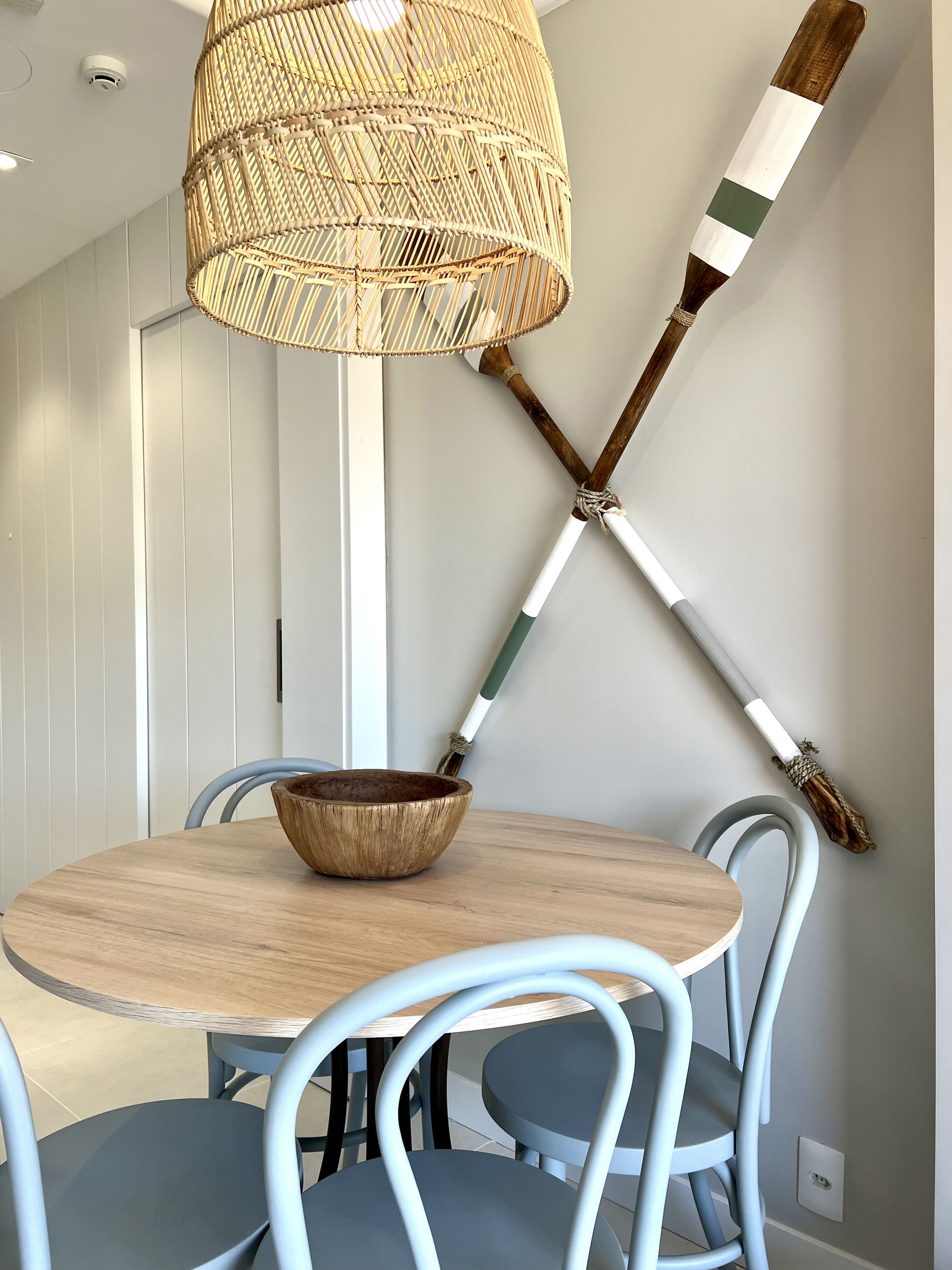 A dining area with a round wooden table, blue chairs, a woven rattan pendant light, and two wooden-handled paintbrushes mounted on the wall.