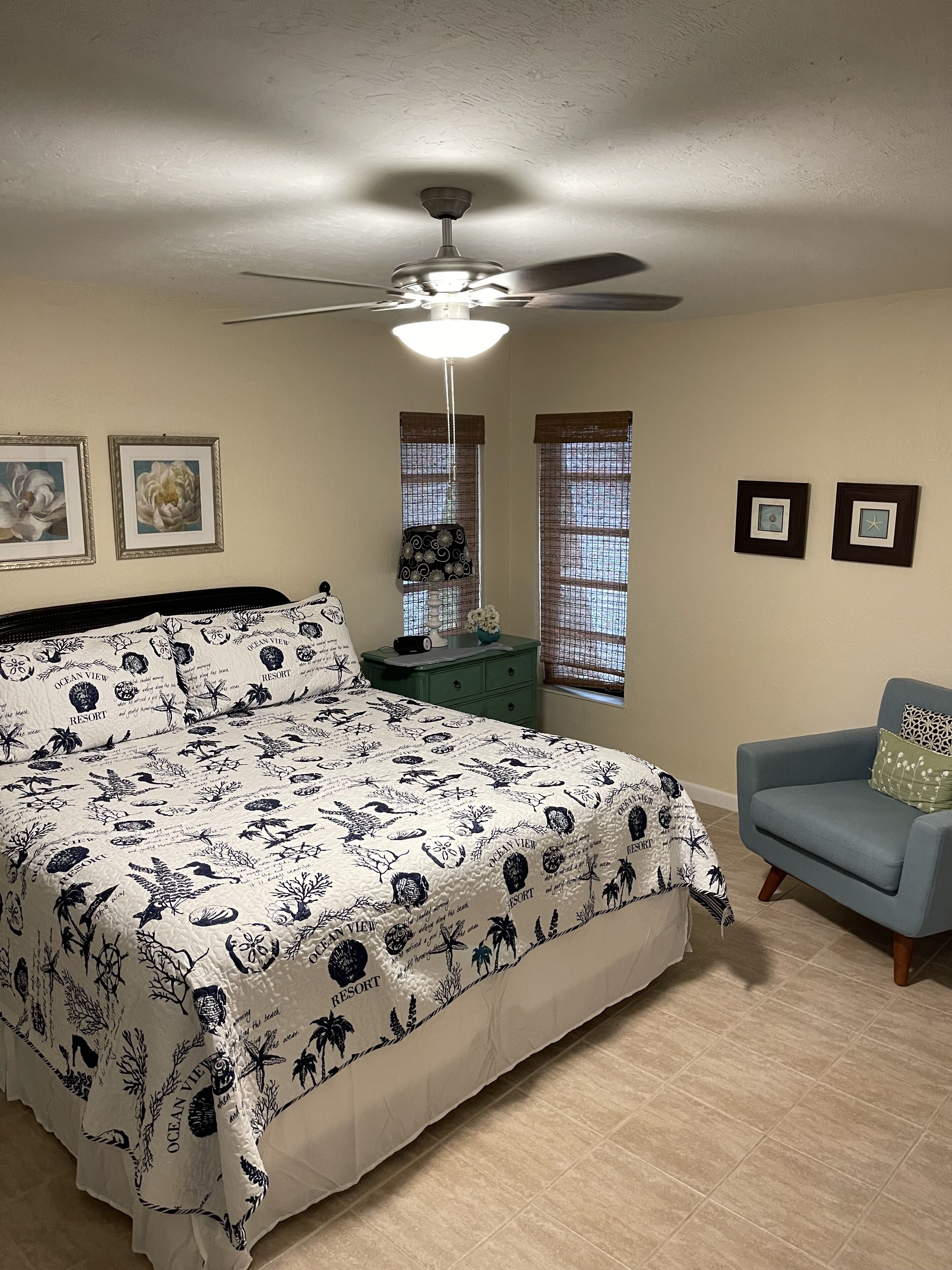 Un dormitorio con cama, ventilador de techo, ventana con vistas y una pequeña mesa con un objeto decorativo.