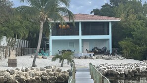 A two-story building with a red-tiled roof, a wooden dock leading to a sandy area with rocks, and palm trees. - 🌊 Private Oasis With Pool Spa Dock and Ocean right in your backyard 🌊 allow pets (Marathon)