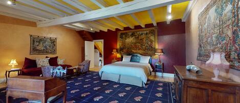 A bedroom with a large bed, a chair, a desk, and a tapestry on the wall.