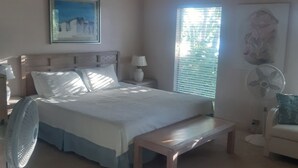 A bedroom with a bed, bedside table, lamp, fan, and artwork on the wall. - Private Island w Causeway to Town, 2 Bedroom, 2 bathroom, Full kitchen, Sleeps 6 (George Town)