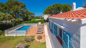 Un área de piscina con valla blanca, sillones de descanso y un edificio con contraventanas azules.