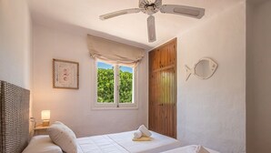 Un dormitorio con dos camas, un ventilador de techo, una ventana con vista a follaje, una puerta de madera y una decoración de peces montada en la pared.