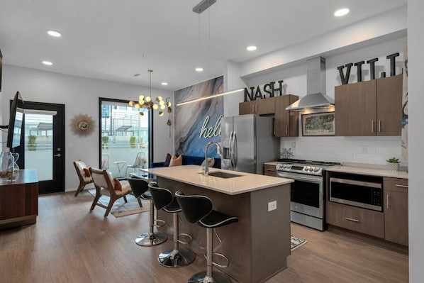 A modern kitchen with a central island, bar stools, a refrigerator, oven, microwave, and a wall-mounted sign with 'NASH' and 'VTT'.