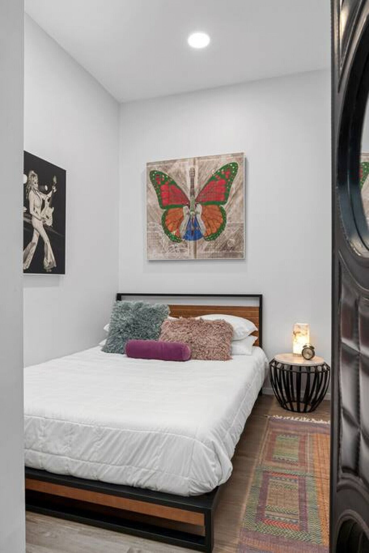 Een slaapkamer met een bed, kussens, een wanddecoratie van een vlinder, een klein nachtkastje met een lamp, en een vloerkleed.