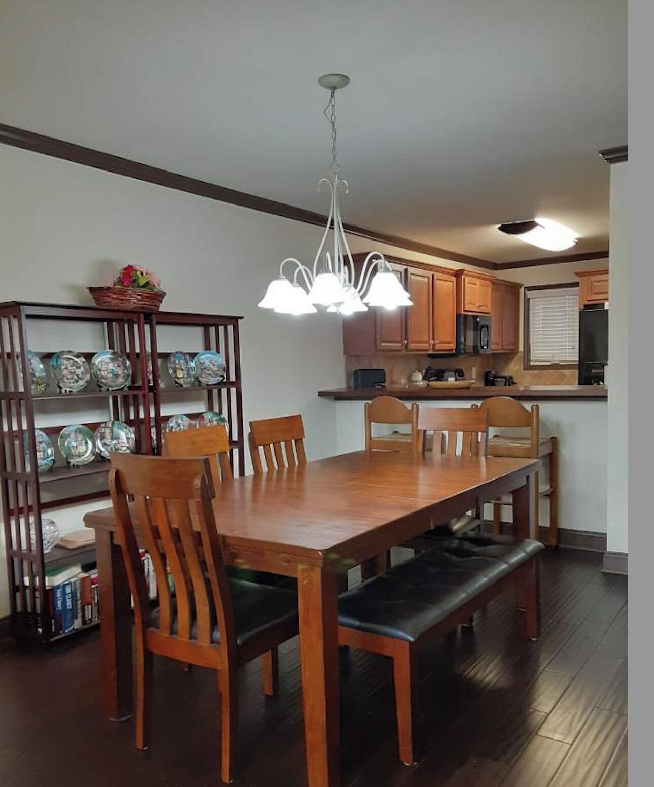 A dining area with a wooden table and chairs, a chandelier, and a kitchen in the background.