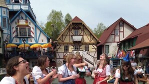 Un groupe de personnes, vêtues de costumes traditionnels, est réuni dans un espace de restauration en plein air, avec des tables et des bancs en bois, le tout étant entouré de bâtiments de style architectural européen.