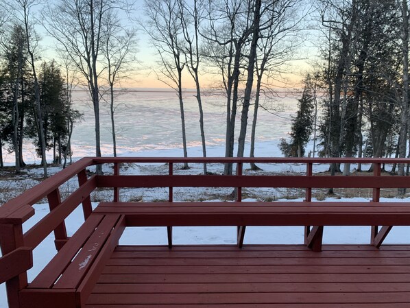 A wooden deck with a metal railing overlooking a snowy landscape with trees and a body of water.