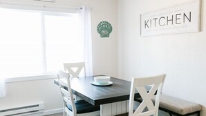 A dining area with a dark wooden table, white chairs, and a bench. A sign on the wall reads "Welcome to the Kitchen." - Cozy 2 Bedroom with Full Kitchen One Block from the Ocean!! (Crescent City)