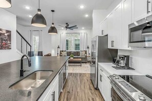 A modern kitchen with a central island, stainless steel appliances, pendant lighting, and a view to a living area with a sofa and curtains. - Highland Station • Rooftop Views • Sleeps 14 (Nashville)