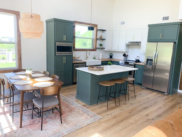 Une cuisine moderne avec des armoires de couleur turquoise, un îlot central doté de tabourets, un réfrigérateur en acier inoxydable et un coin repas avec une table et des chaises en bois.