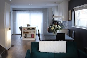 A modern living room with a green sofa, a dining area with a table and chairs, and a kitchen with cabinets and a window.