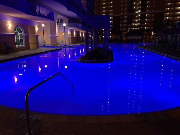 Une piscine intérieure dont la surface de l’eau est illuminée de lumière bleue, entourée d’un bâtiment doté de nombreux balcons et d’un espace couvert aménagé avec des sièges.