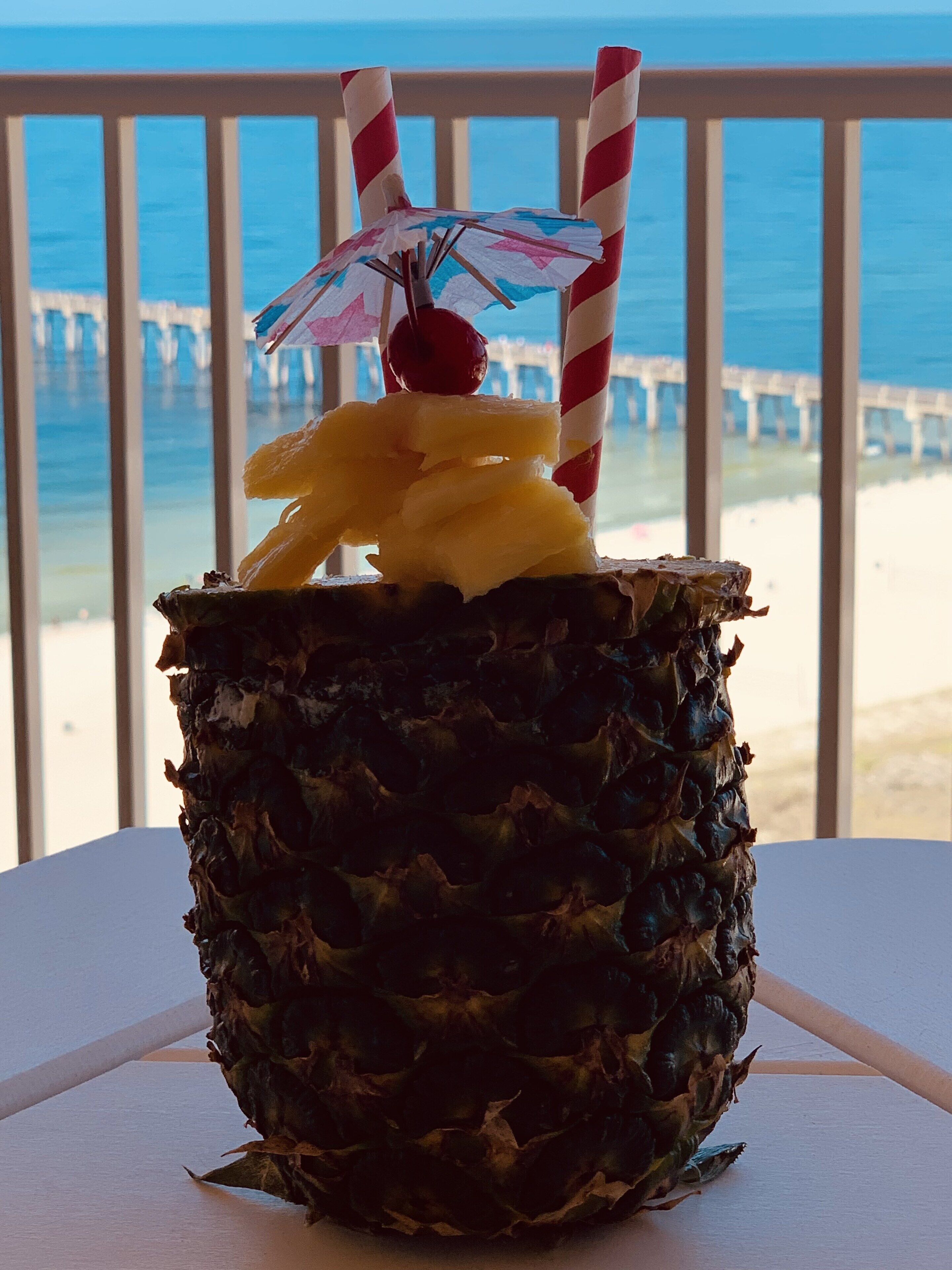 Un cocktail tropical servi dans une demi-ananas, avec une paille et un petit parasol, le tout présenté sur fond de vue sur l’océan et une jetée.