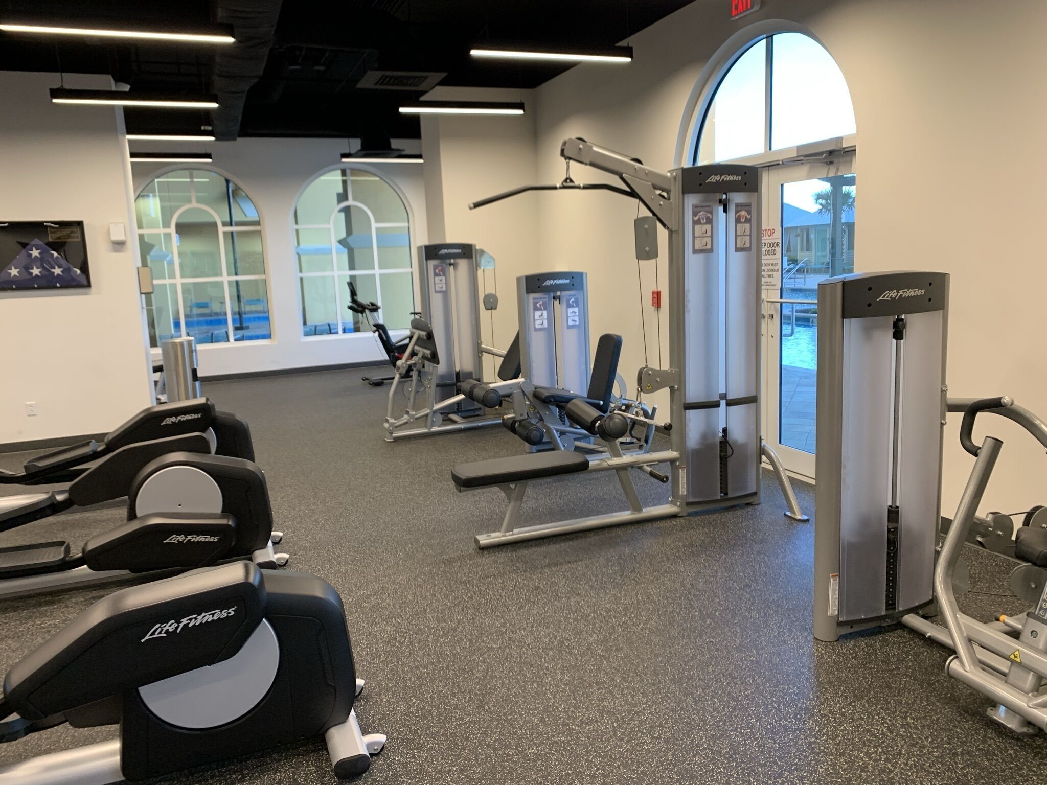 Un gimnasio con diversas máquinas de ejercicio, un ventanal grande y una puerta con la leyenda 'STOP DOOR CLOSED'.