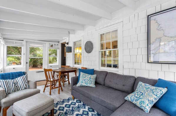 A cozy living room with a grey sofa, blue patterned pillows, a wooden dining table, and white brick walls. - Historic Cottage on White Pond, near Downtown Chatham and beaches (Chatham)