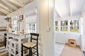A kitchen with white cabinets, a wooden table, and a chair. There is a window with a view of trees and a basket on the floor. - Historic Cottage on White Pond, near Downtown Chatham and beaches (Chatham)