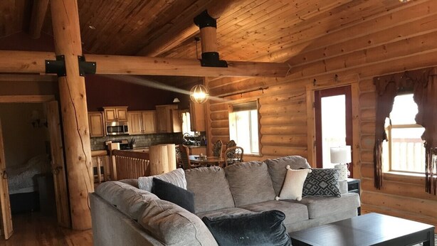 A log cabin interior with a living room featuring a gray sectional sofa, a coffee table, and a kitchen area in the background. - Log Home on the Blackfoot River (Lincoln)