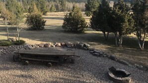 A rustic outdoor setting with a wooden bench, a fire pit, and a gravel area surrounded by trees and mountains in the background. - Log Home on the Blackfoot River (Lincoln)