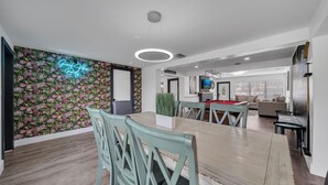Ein moderner Essbereich mit Holztisch und hellgrünen Stühlen, einem Neon-Leuchtschild an der Wand und Blick in einen Wohnraum mit Sofa und Fernseher.