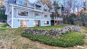 A two-story house with a stone wall and a wooden deck. - Oceanfront, views from every room! Sunny, spacious midcoast retreat (Round Pond)