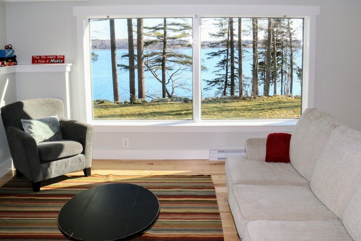 Un salon doté d’une grande fenêtre donnant sur un lac et des arbres, d’un fauteuil gris, d’un canapé blanc et d’un tapis à rayures.