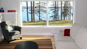 A living room with a large window overlooking a lake and trees, a gray armchair, a white sofa, and a striped rug. - Oceanfront, views from every room! Sunny, spacious midcoast retreat (Round Pond)