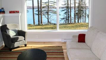 Un salon doté d’une grande fenêtre donnant sur un lac et des arbres, d’un fauteuil gris, d’un canapé blanc et d’un tapis à rayures.