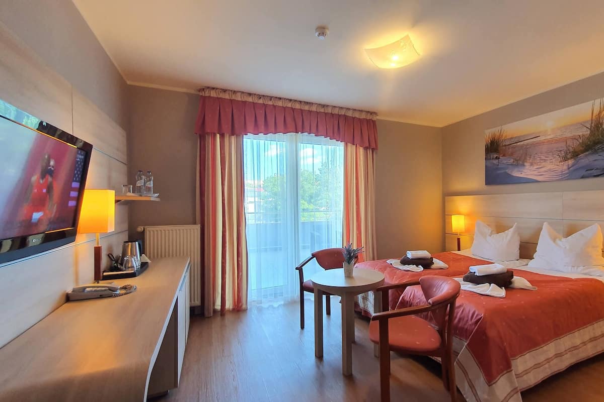 Ein Hotelzimmer mit einem Bett, zwei Nachttischen mit Lampen, einem Fernseher auf einem Schrank, einem Sessel, einem kleinen Tisch mit einer Pflanze und einem Fenster mit Vorhängen.